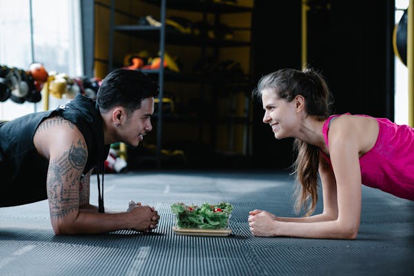 A man and a woman plunking in front of each other, with a leafy vegetable and fruits in the middle of them.
